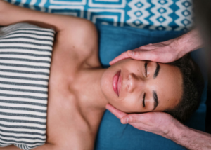 A woman lying on a massage table with her eyes closed, receiving a gentle facial massage in a peaceful, relaxing setting